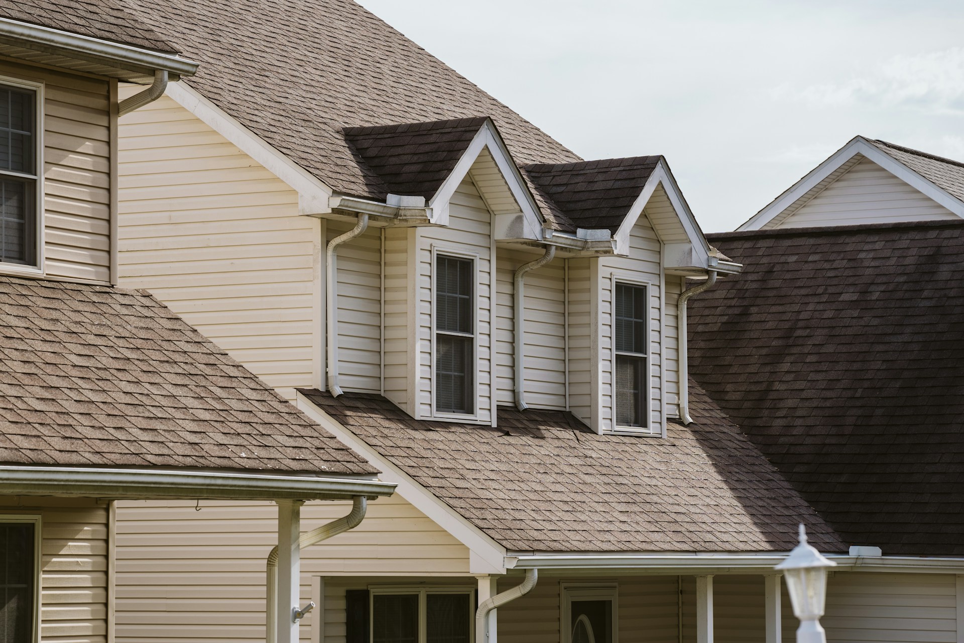 Close-up of a beige suburban house roof with dormer windows, white trim and gutters against a blue sky.