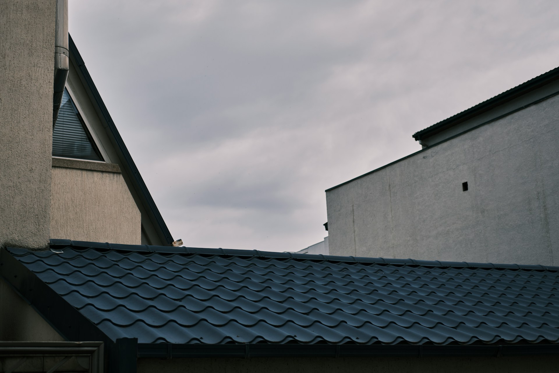 Metal Roofing | RAINTITE Roofing & Construction Two neighboring buildings with a blue, wavy-tiled roof in the foreground and a cloudy sky above.
